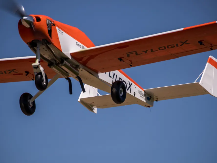 A small orange and white Flylogix aircraft with landing gear extended flies against a clear blue sky, its propeller spinning in motion.