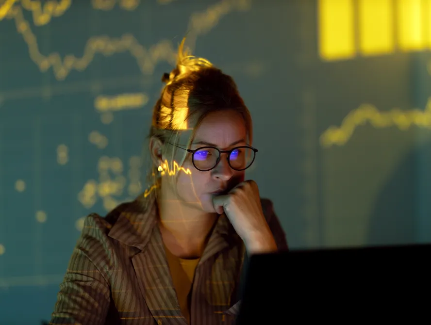 Focused woman in glasses working on a laptop at home with stock market numbers and charts projected on her face.