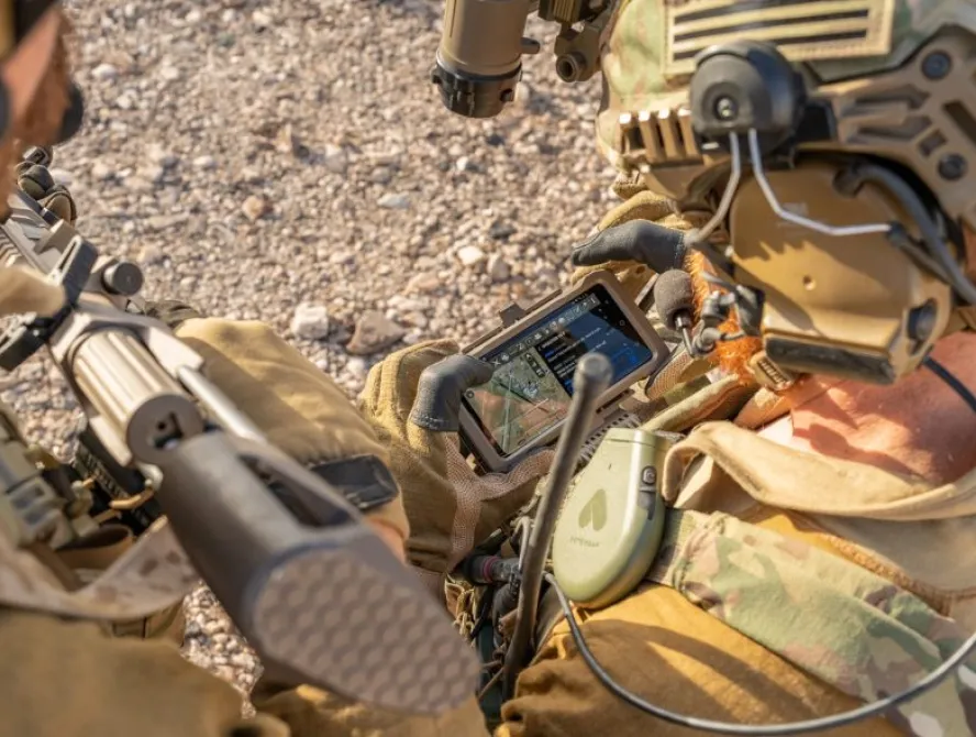 Two soldiers in tactical gear view a rugged handheld device displaying a satellite map while coordinating in a rocky outdoor environment.