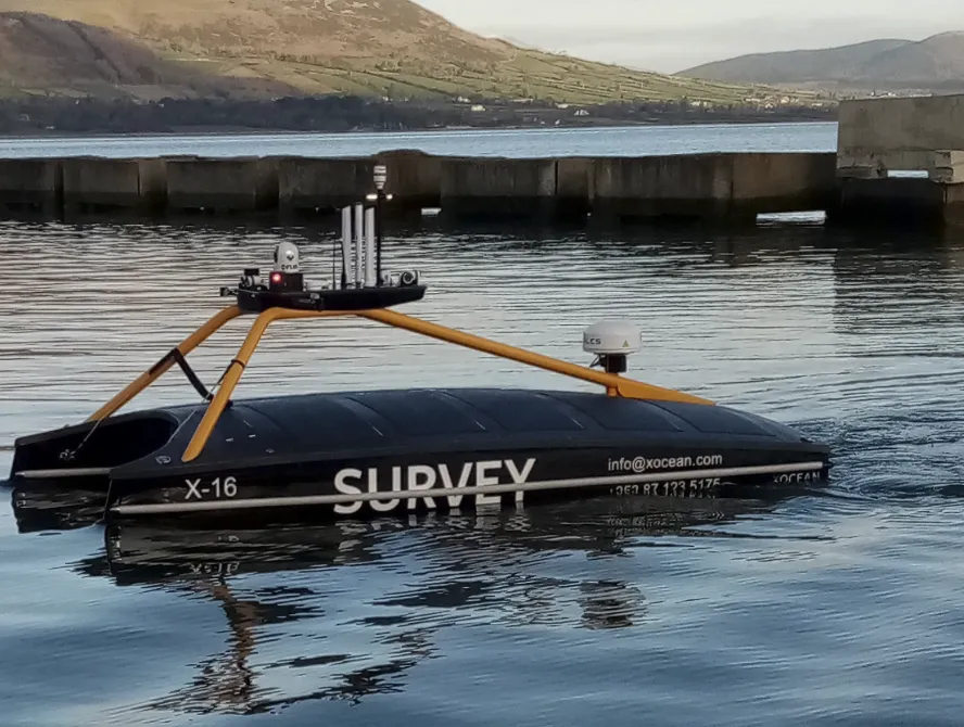 Black and yellow marine survey vessel labeled ‘SURVEY’ floating on calm water near a concrete dock