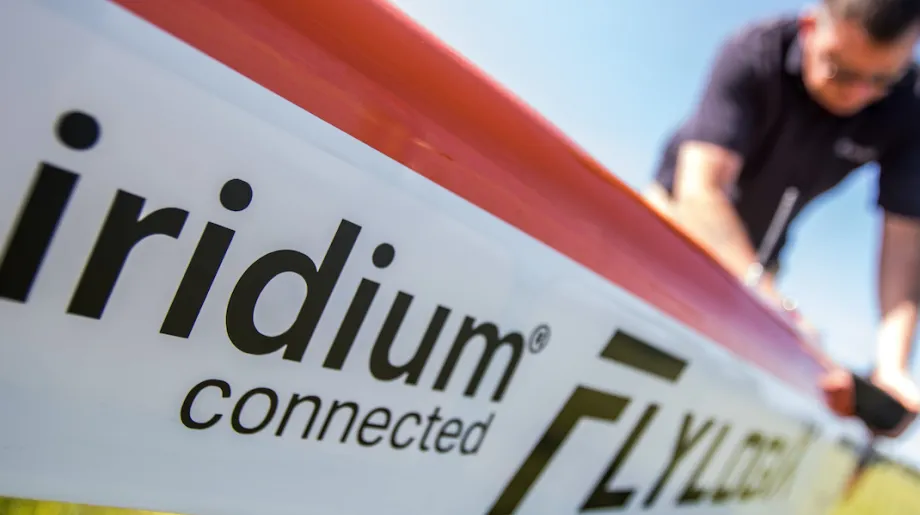 Close-up of an aircraft fuselage labeled “Iridium connected Flylogix,” with a person in the background working on the plane outdoors under a clear blue sky.