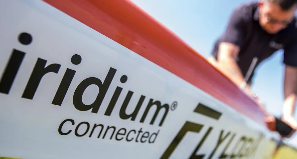 Close-up of an aircraft fuselage labeled “Iridium connected Flylogix,” with a person in the background working on the plane outdoors under a clear blue sky.