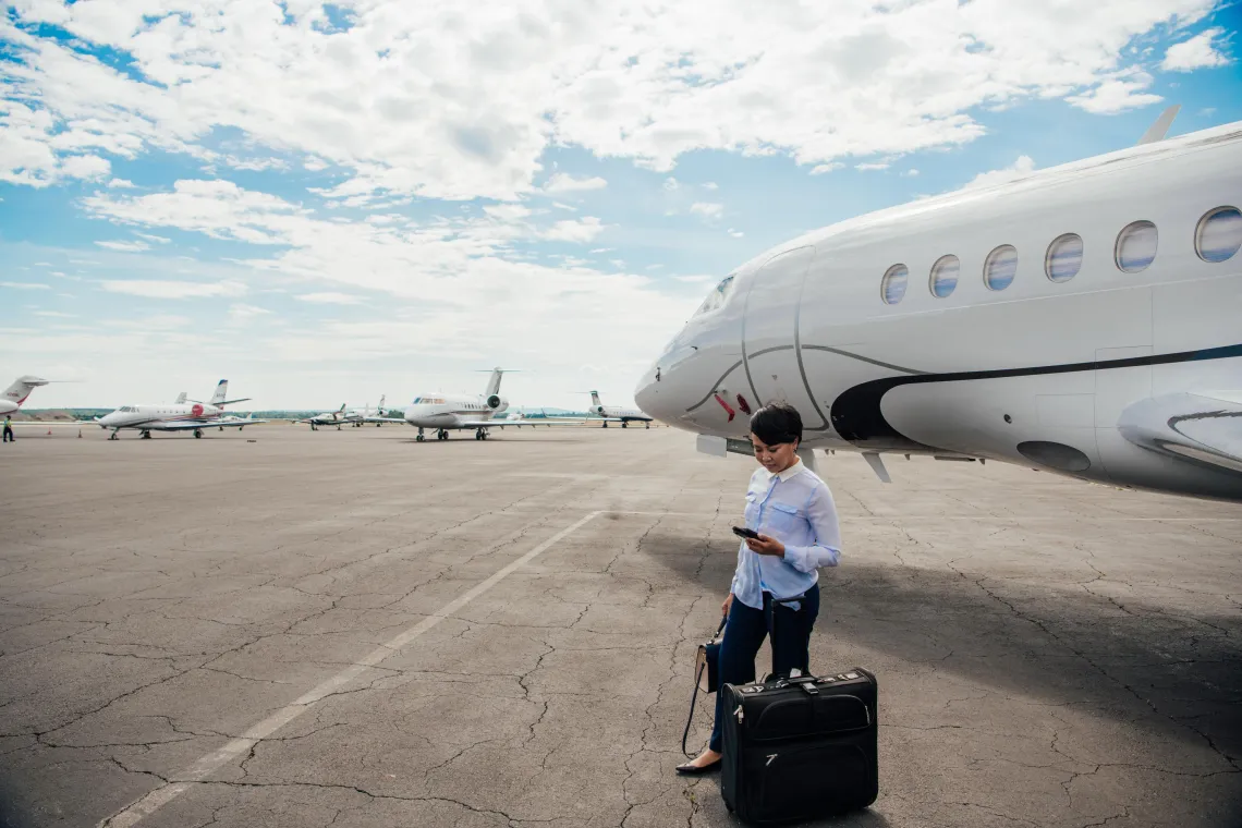 A woman stands on an airport tarmac beside a private jet, holding her phone and suitcase, with several other small aircraft parked in the background under a partly cloudy sky.