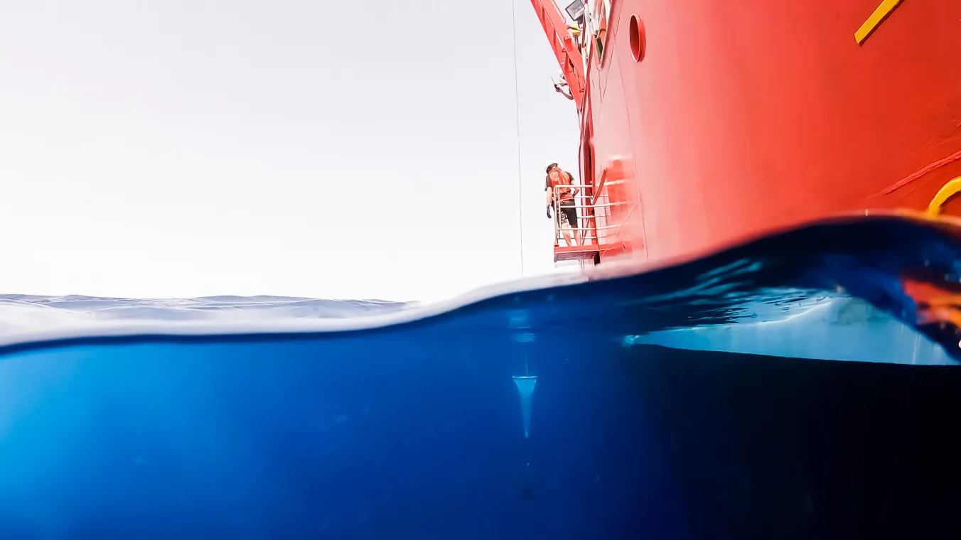 A red research ship sits on the ocean surface with equipment submerged beneath calm waters as marine scientists prepare for oceanographic study