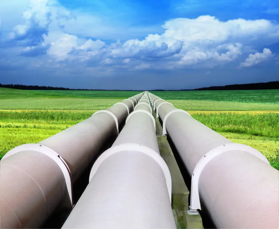 Large metal pipelines stretch into the distance across a green grassy field under a bright blue sky with scattered white clouds.