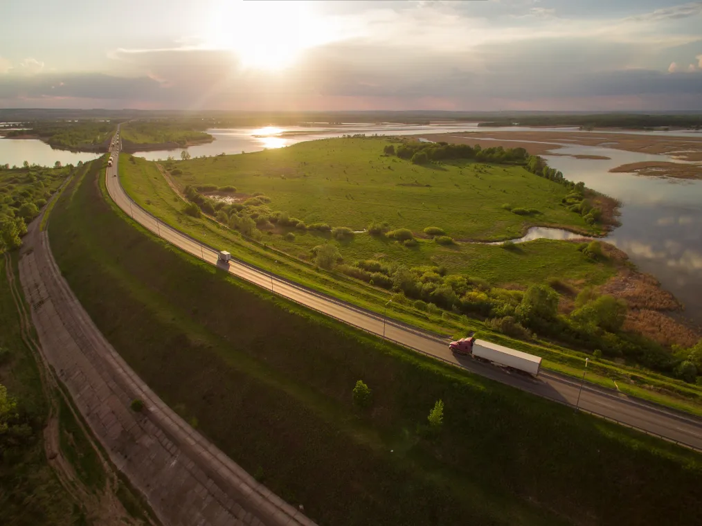 A red semi-truck driving along a winding highway beside green fields and water at sunset, with golden light reflecting off the landscape and distant horizon.