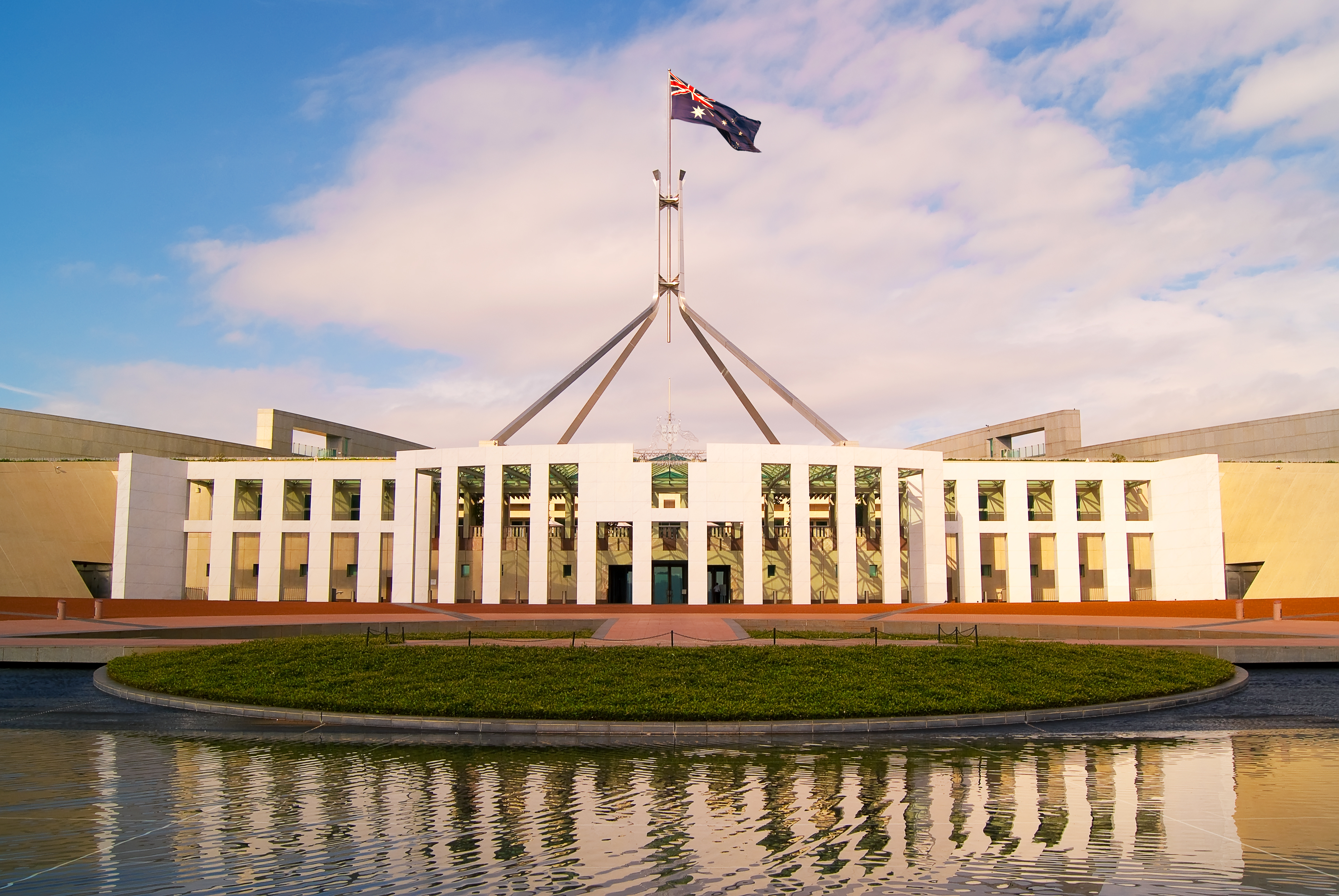 The Australian Parliament House with a large flag atop its spire reflected in a calm pool.