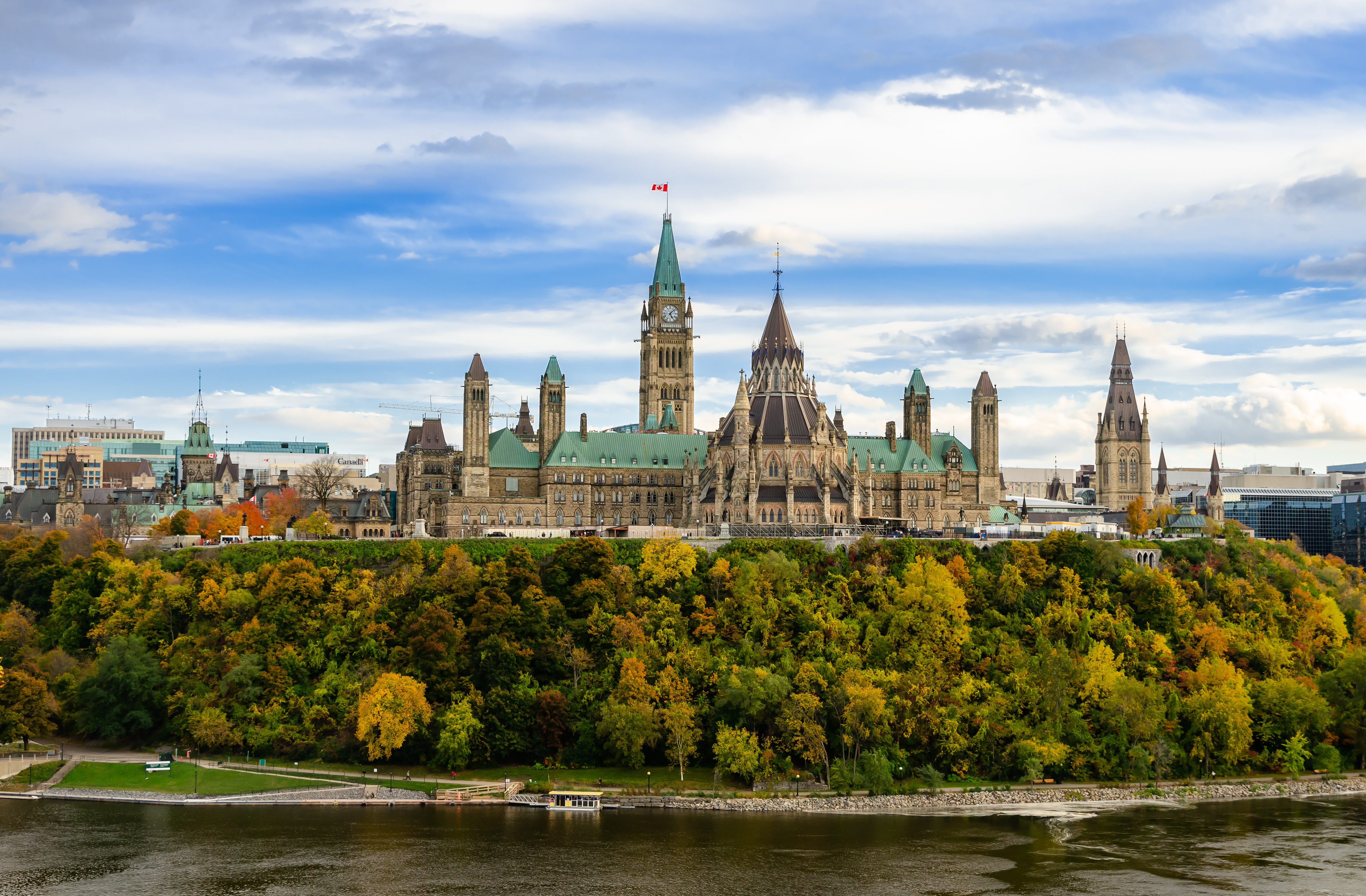 Canada’s Parliament Buildings sit atop a hill surrounded by fall trees overlooking the Ottawa River.