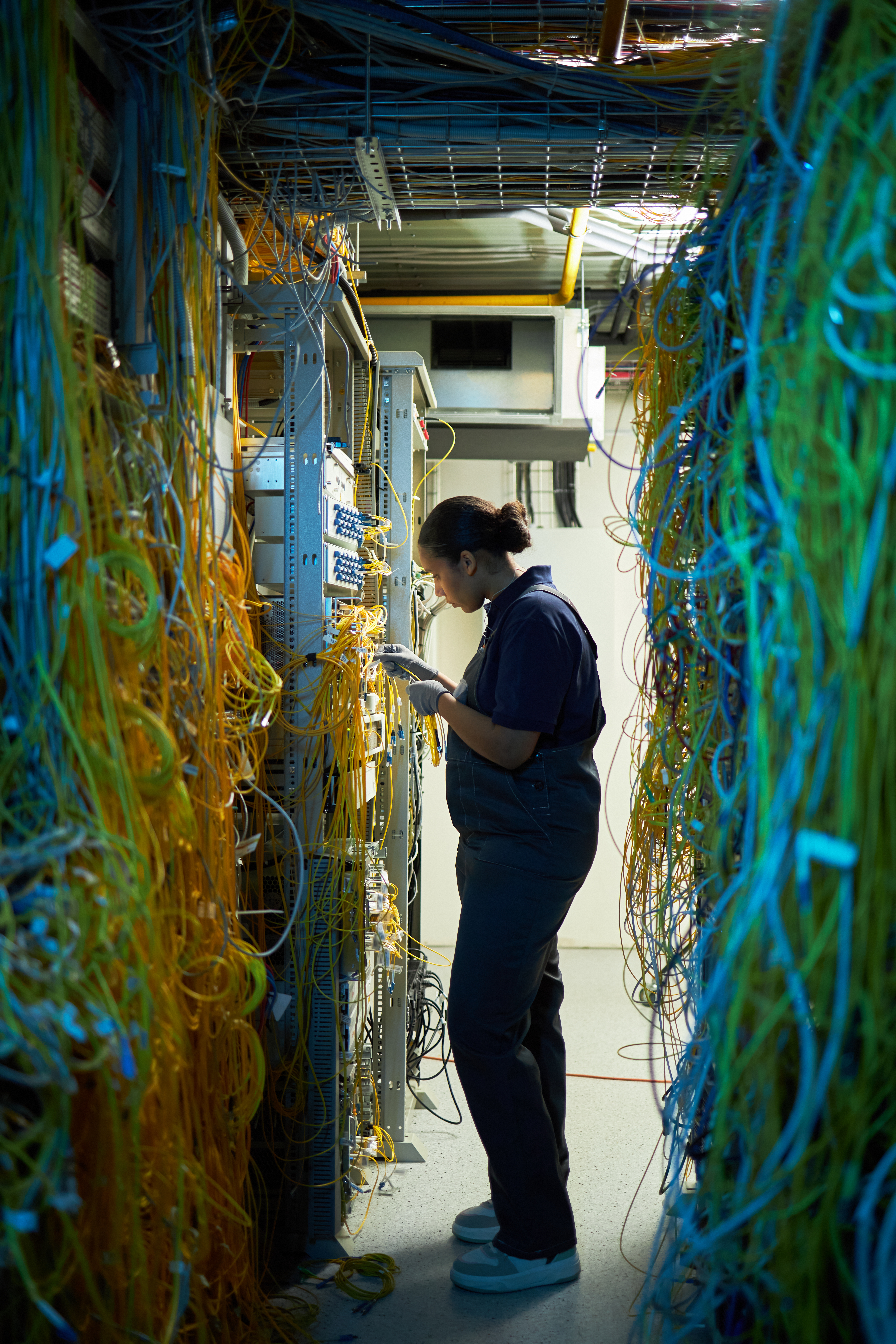A female technician wearing gloves works on a piece of networking hardware, standing between two walls of yellow and blue data cables in a server room.