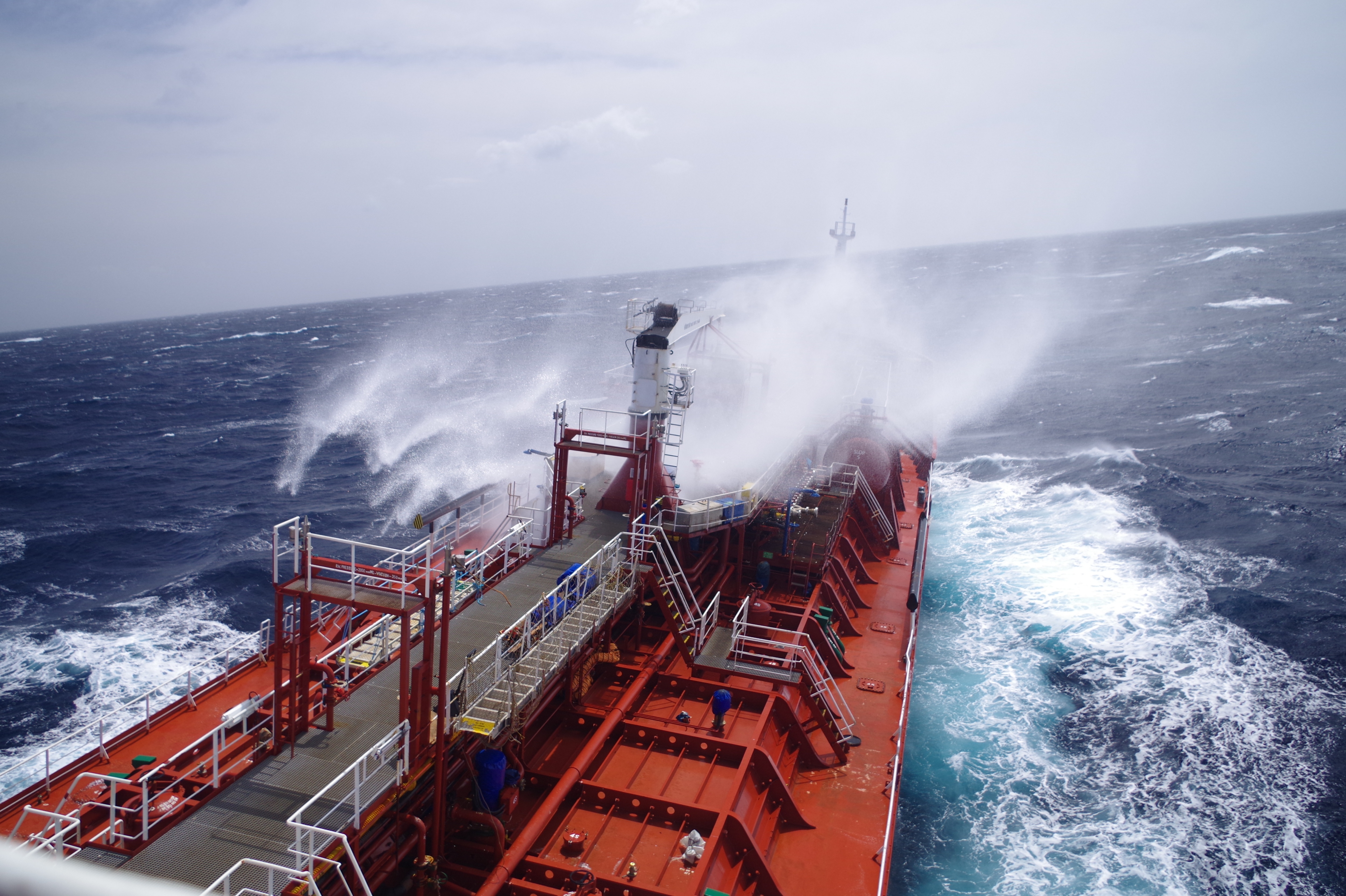 A large, red, chemical tanker ship sails through rough, stormy seas with waves crashing over the bow.