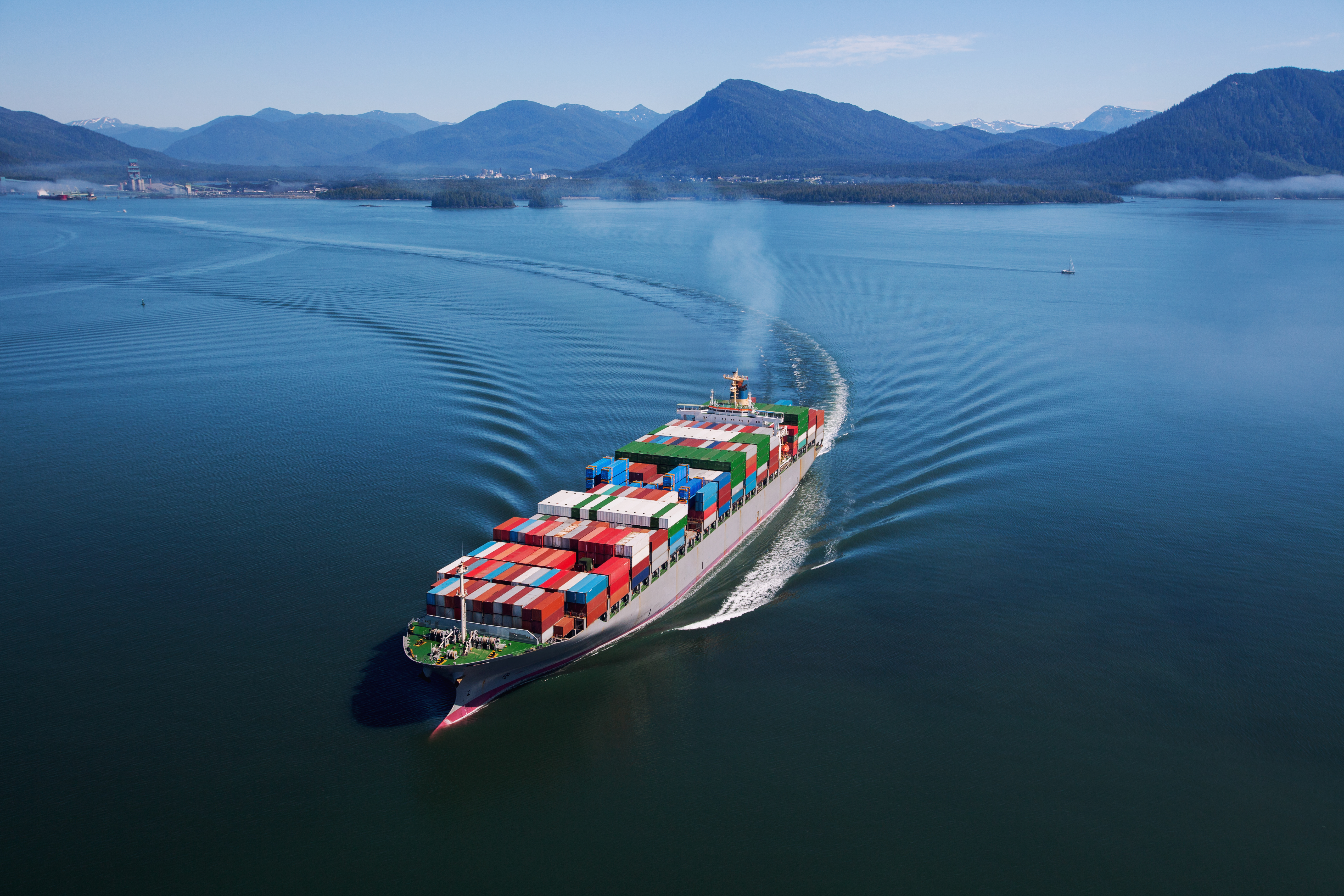 A cargo ship loaded with colorful containers sails through calm blue waters surrounded by mountains.