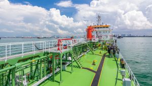 View from the deck of a brightly colored oil tanker, with many other ships visible in a busy port and city skyline under a sunny sky.