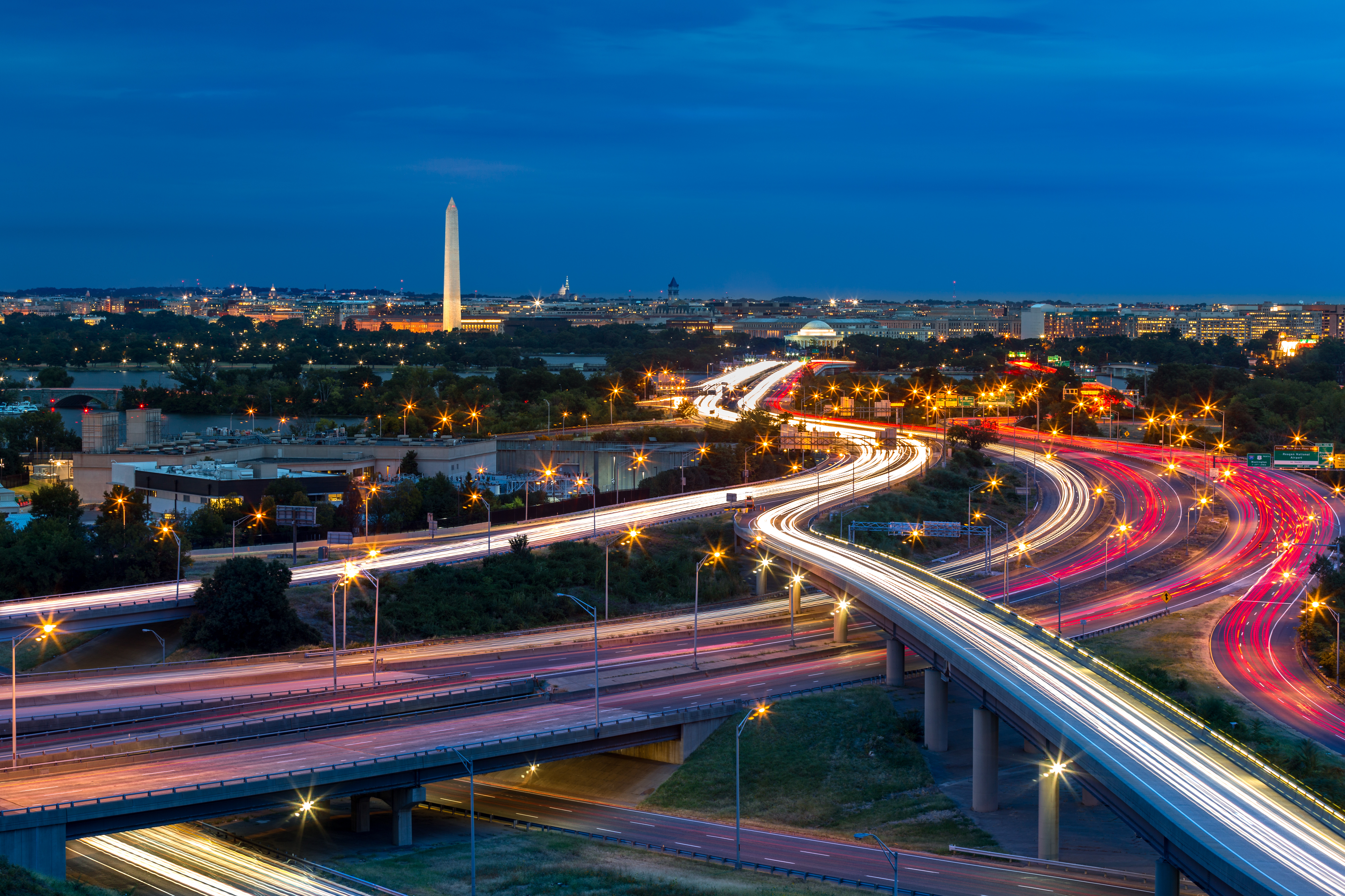 Curving highways with light trails lead toward the Washington Monument at dusk.