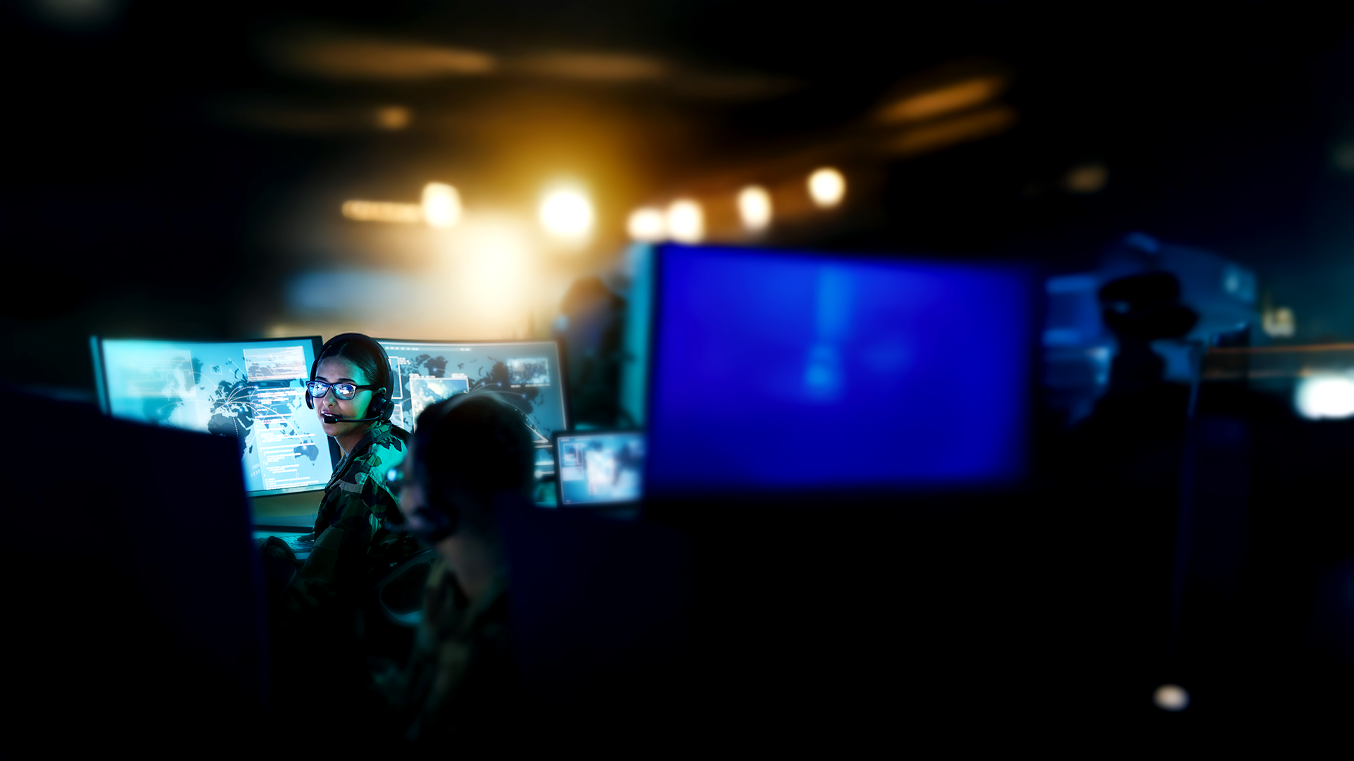 A man and a woman in military uniforms, wearing headsets, work in a dimly lit control room, monitoring digital maps and data on multiple screens.